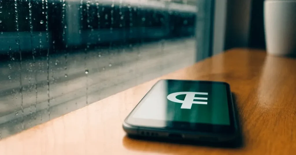 Ein Café-Tisch am Fenster trägt ein Smartphone mit dunklem Display, an der Scheibe laufen Regentropfen herunter und draussen steht eine Tram.