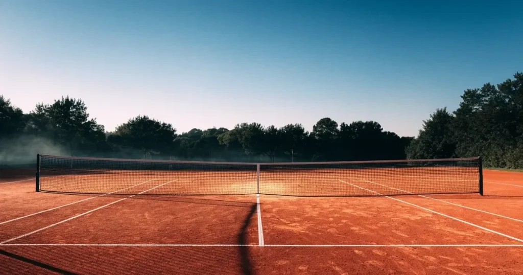 Ein Tennisplatz aus Sand bei Sonnenaufgang mit gespanntem Netz, Nebel zwischen den Bäumen am Rand