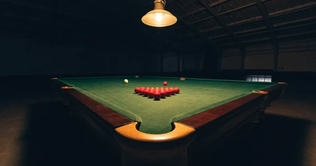 Ein Snookertisch mit grünem Tuch und einem Dreieck aus roten Kugeln unter einer tief hängenden Lampe in einer leeren Halle.