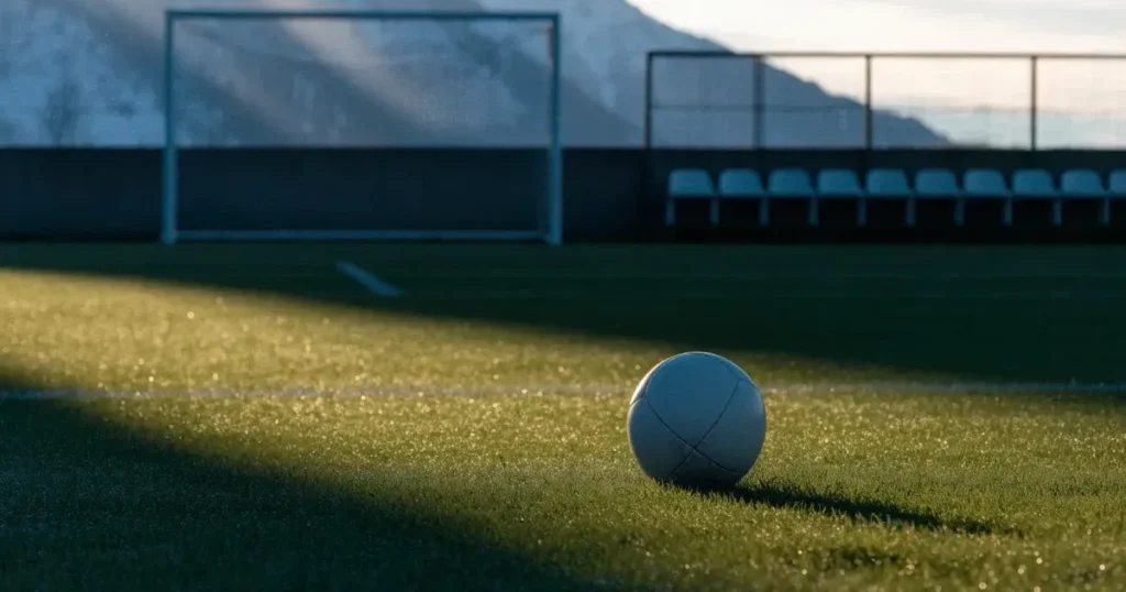 Ein Fussball liegt am frühen Morgen auf dem Rasen eines leeren Fussballfeldes, dahinter eine kleine Tribüne und schneebedeckte Berge.